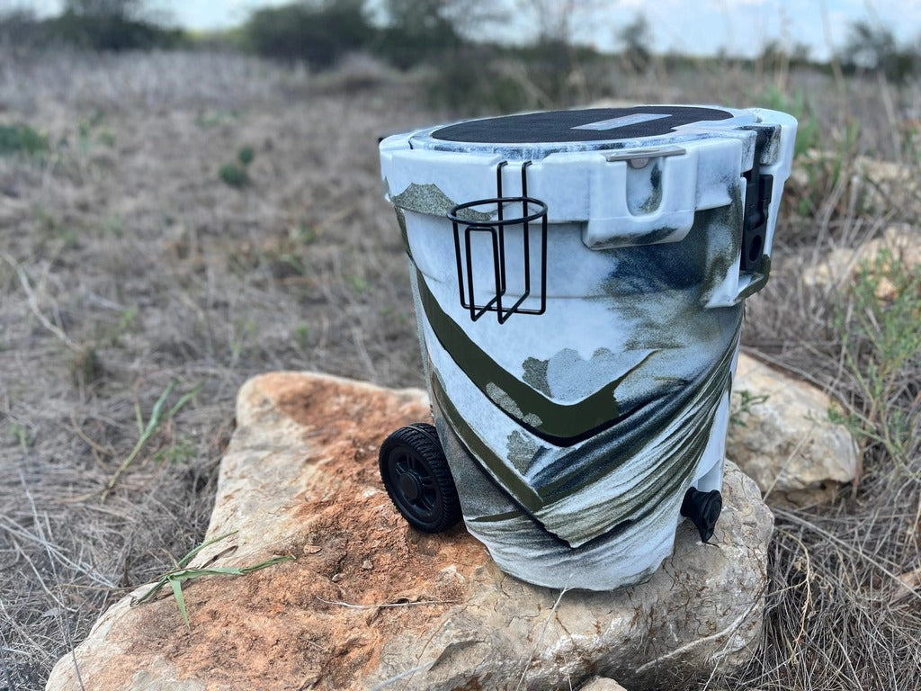 Iceland Coolers 7.5-gal camo cooler with cup holder, wheels, and drain spout, resting on a rock in dry grass under open sky.