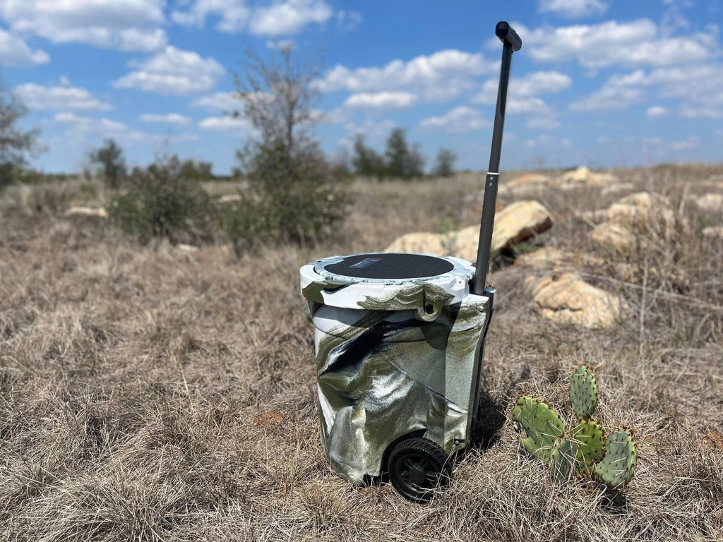 Iceland Coolers 7.5-gal camo cooler with wheels and handle, shown in dry, grassy terrain—built for mobility, hunting, and rugged use.