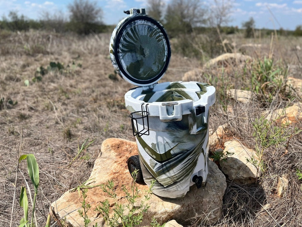 Iceland Coolers 7.5-gal camo cooler with lid open, cup holder on side, and rear wheels, sitting on rocks in a dry grassy field.