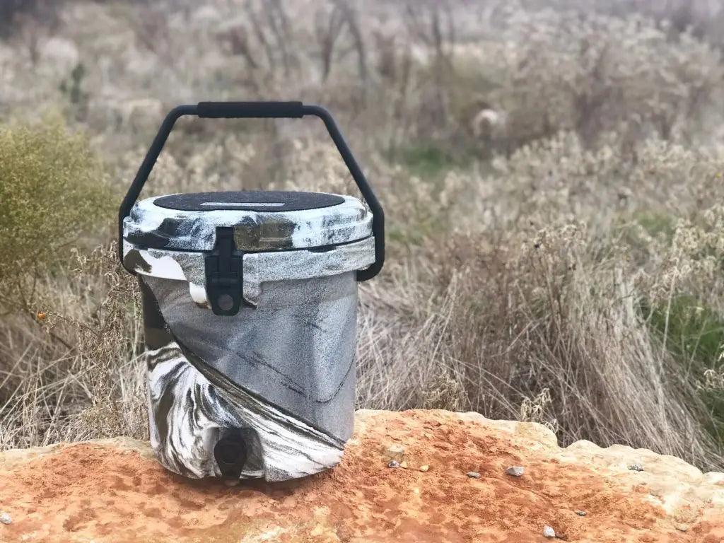 Iceland Coolers 5-gal marbled bucket cooler with spout and carry handle, sitting on a rock in dry grass—rugged, rotomolded design.