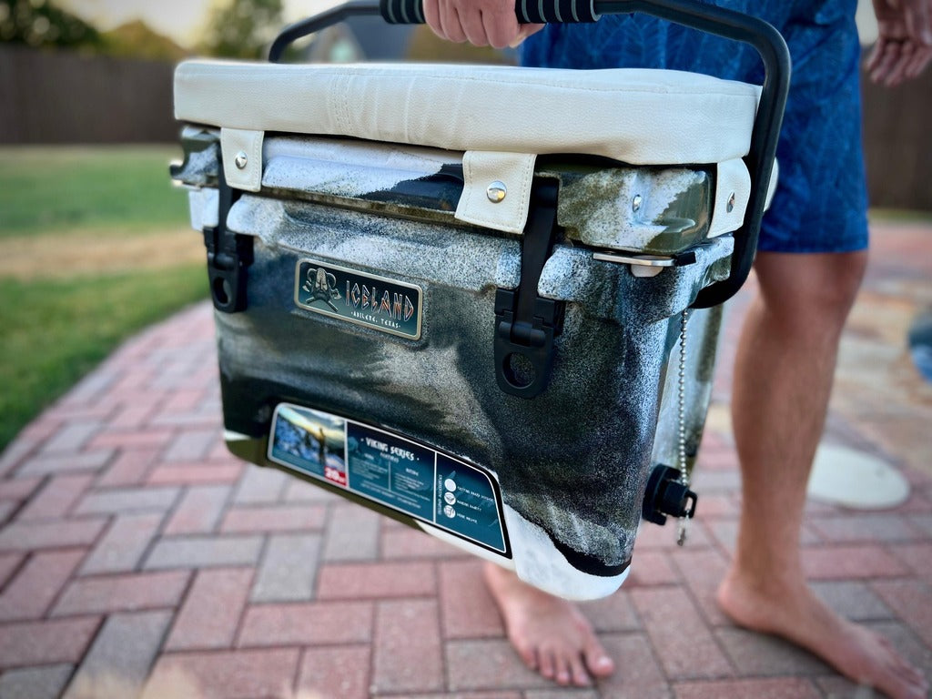Person carrying camo Iceland Cooler with padded white seat cushion, designed for comfort, portability, and sunlight reflection.