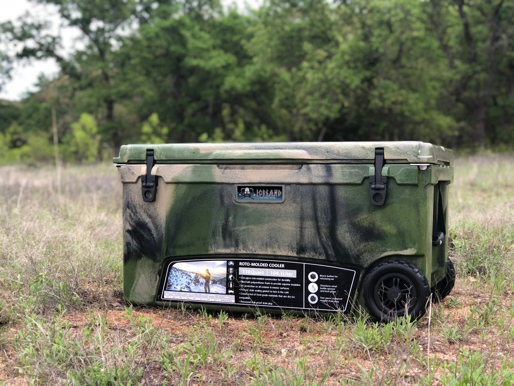 Iceland Coolers Longship Series 70-quart roto-molded green camo cooler with wheels, shown outdoors in a grassy field.