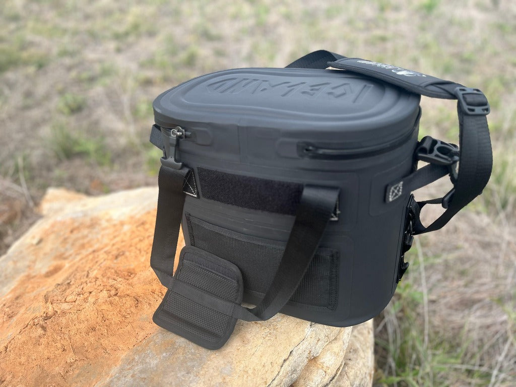 Back view of Iceland Coolers Saga Series 12-can soft cooler in black with padded strap, placed on a rock in a grassy outdoor setting.