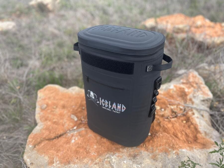 Black Iceland Coolers Saga Series 30-can soft cooler with side loops and front zipper pocket, shown outdoors on a large rock.