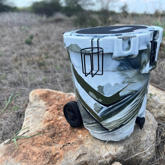 Iceland Coolers 7.5-gal camo cooler with cup holder, wheels, and drain spout, resting on a rock in dry grass under open sky.