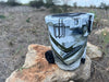 Iceland Coolers 7.5-gal camo cooler with cup holder, wheels, and drain spout, resting on a rock in dry grass under open sky.