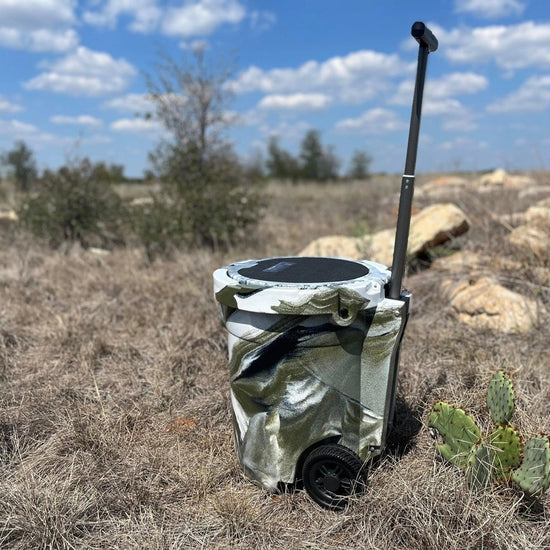 Iceland Coolers 7.5-gal camo cooler with wheels and handle, shown in dry, grassy terrain—built for mobility, hunting, and rugged use.