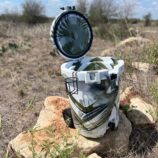 Iceland Coolers 7.5-gal camo cooler with lid open, cup holder on side, and rear wheels, sitting on rocks in a dry grassy field.