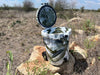 Iceland Coolers 7.5-gal camo cooler with lid open, cup holder on side, and rear wheels, sitting on rocks in a dry grassy field.