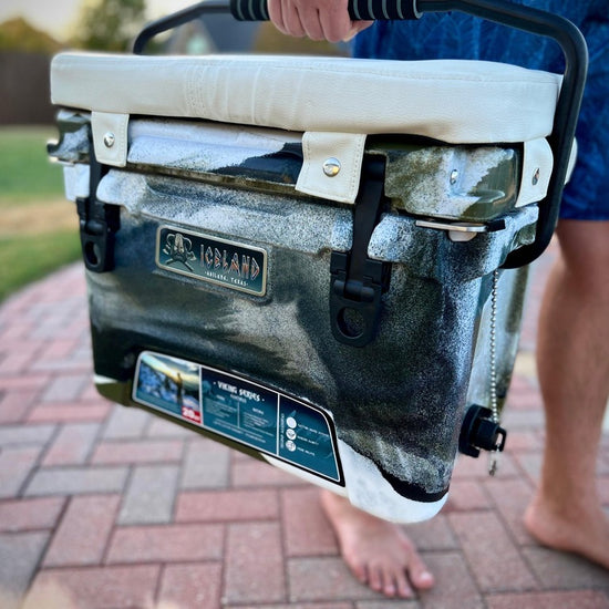 Person carrying camo Iceland Cooler with padded white seat cushion, designed for comfort, portability, and sunlight reflection.