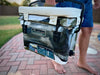 Person carrying camo Iceland Cooler with padded white seat cushion, designed for comfort, portability, and sunlight reflection.