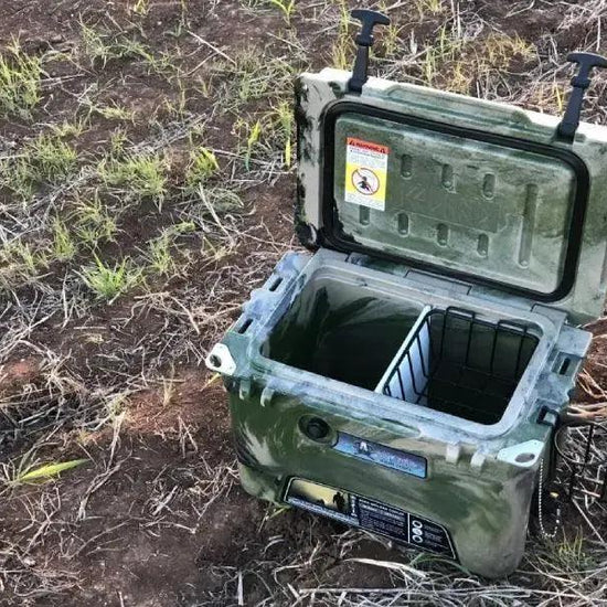 Iceland Coolers camo ice chest with cutting board divider and wire basket, open outdoors on grass and dirt terrain.
