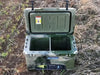 Open green camo Iceland Cooler outdoors, featuring a center divider and black hanging basket for dry storage during hunting or camping.