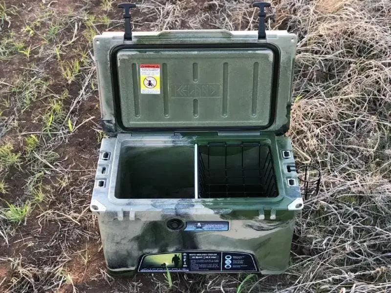 Open green camo Iceland Cooler outdoors, featuring a center divider and black hanging basket for dry storage during hunting or camping.