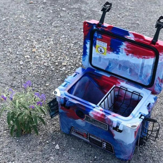 Open Iceland Cooler in red, white, and blue camo with hanging cup holder and interior basket, shown outdoors on gravel near wildflowers.