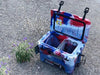 Open Iceland Cooler in red, white, and blue camo with hanging cup holder and interior basket, shown outdoors on gravel near wildflowers.