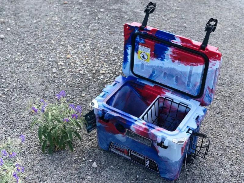Open Iceland Cooler in red, white, and blue camo with hanging cup holder and interior basket, shown outdoors on gravel near wildflowers.
