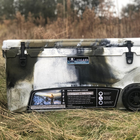 Iceland Coolers Longship Series 110-quart roto-molded wheeled cooler in camo design, shown outdoors in a grassy field.