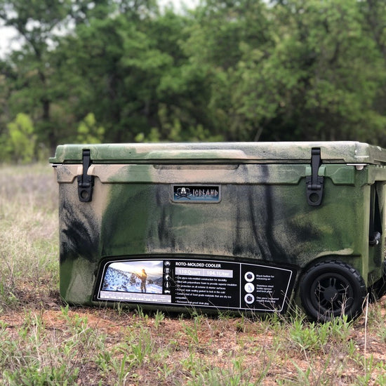 Iceland Coolers Longship Series 70-quart roto-molded green camo cooler with wheels, shown outdoors in a grassy field.