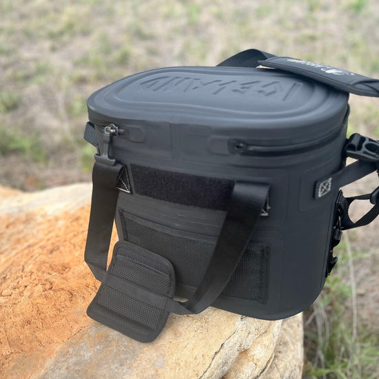 Back view of Iceland Coolers Saga Series 12-can soft cooler in black with padded strap, placed on a rock in a grassy outdoor setting.