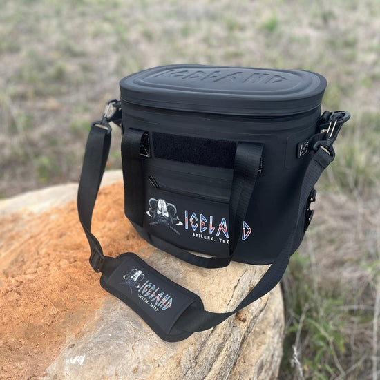 Iceland Coolers Saga Series 12-can soft cooler in black with shoulder strap, shown outdoors on a rock in a grassy field.