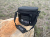 Iceland Coolers Saga Series 12-can soft cooler in black with shoulder strap, shown outdoors on a rock in a grassy field.