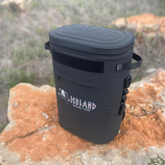 Black Iceland Coolers Saga Series 30-can soft cooler with side loops and front zipper pocket, shown outdoors on a large rock.